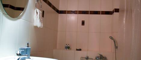Une salle de bain avec un lavabo blanc, un miroir, une douche équipée d’un pommeau de douche à main et une serviette suspendue sur un porte-serviettes.
