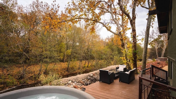 Un jacuzzi en una terraza de madera con mobiliario exterior, rodeado de follaje otoñal.