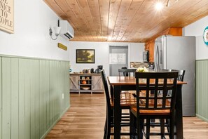 A dining area with a wooden table and chairs, a kitchen in the background, and a green-painted wall.