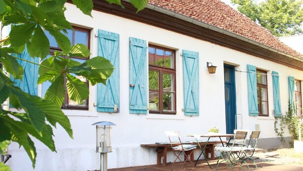 Un bâtiment blanc avec des volets bleus, un toit en tuiles et une terrasse en bois avec une table et des chaises.