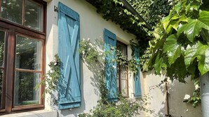 Un bâtiment doté de trois fenêtres, chacune ornée de volets bleus, et entouré de verdure et de plantes en fleurs.