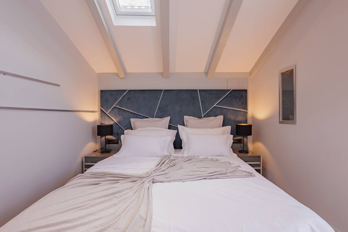 A modern bedroom with a skylight, a bed with white linens, two bedside tables with lamps, and a geometric headboard.