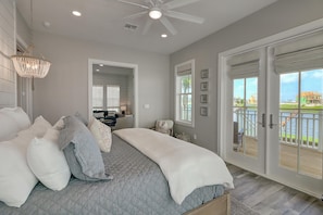 A bedroom with a large bed, a chandelier, a ceiling fan, and a view of a body of water through a sliding glass door. - Seas Serenity 218 LAK (Port Aransas)