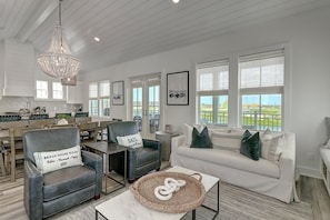 A spacious living room with a white sofa, two armchairs, a coffee table, and a chandelier. There are large windows with a view of a green landscape. - Seas Serenity 218 LAK (Port Aransas)
