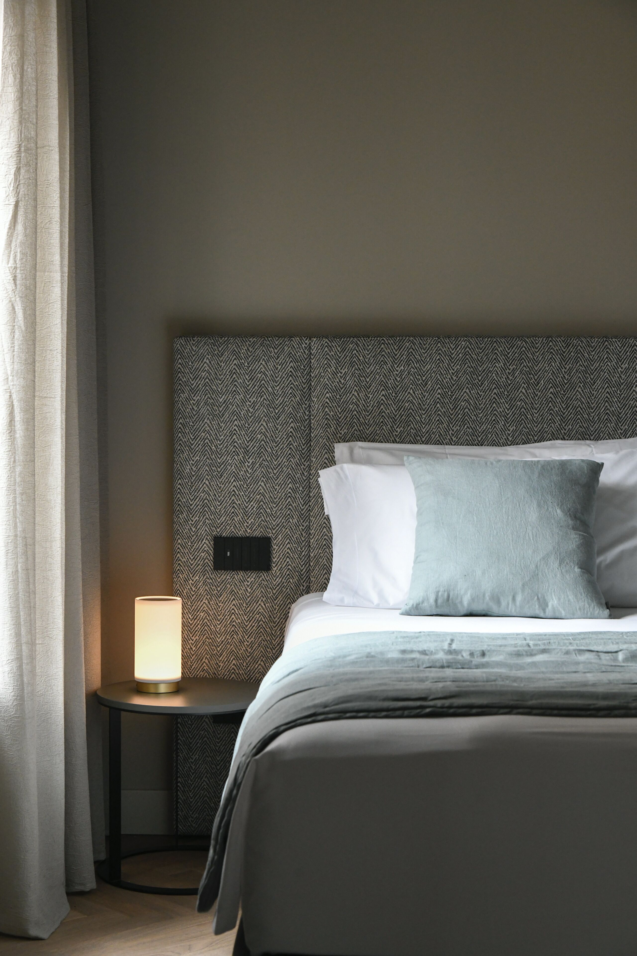 Un letto ben rifatto con una testiera decorata, una lampada da comodino e un piccolo tavolino.