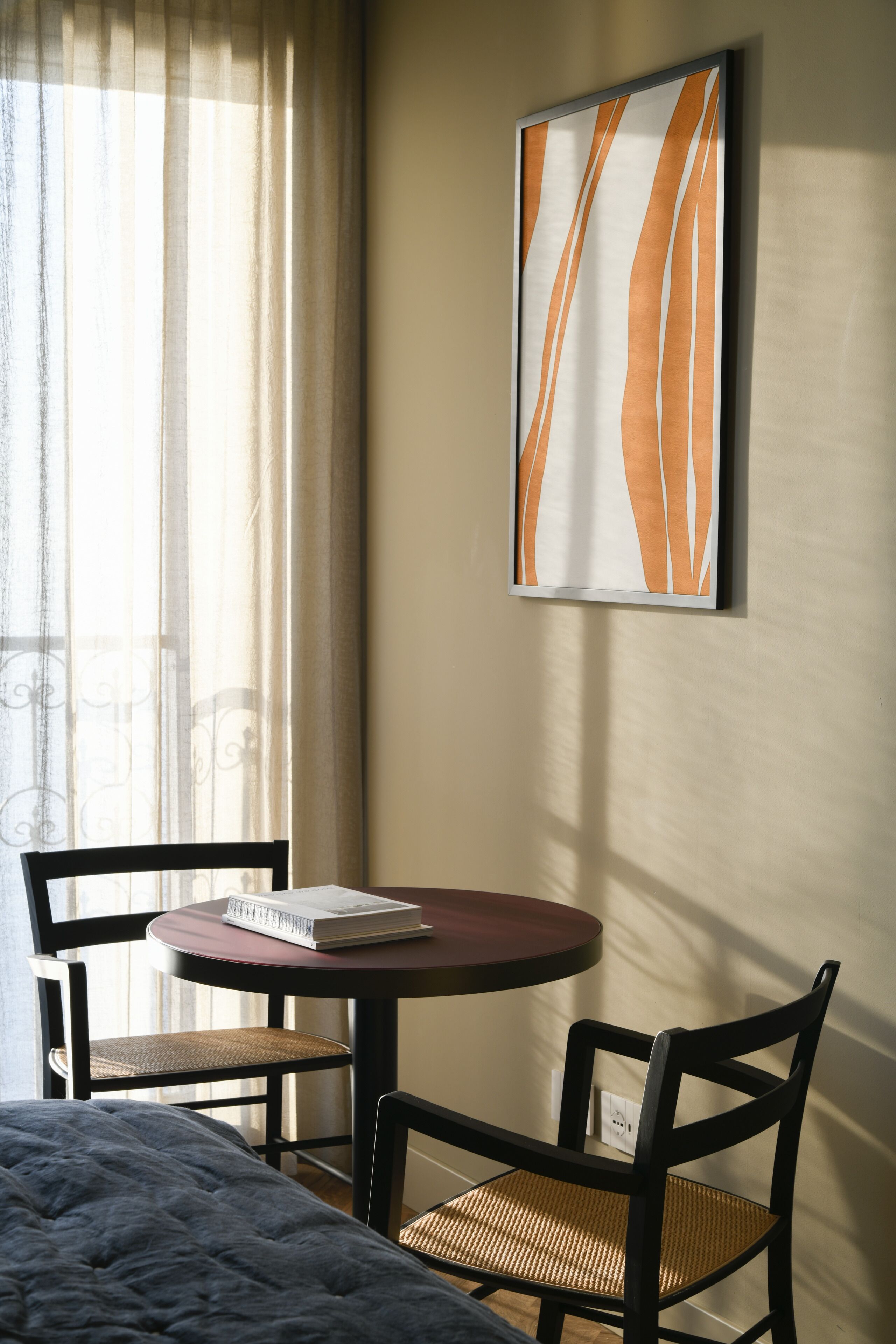 Quarto de hotel com cama, mesa redonda com um livro, duas cadeiras e uma obra de arte abstrata emoldurada na parede.