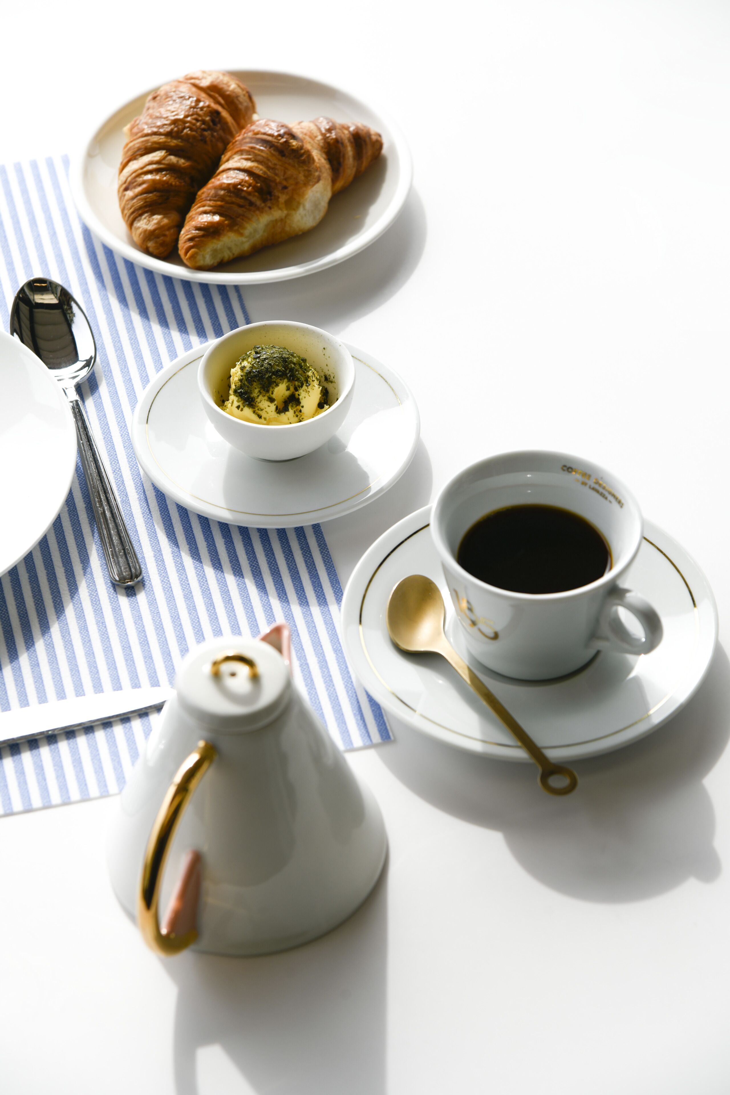 A table set with croissants, a cup of coffee, a bowl of butter, a teapot, and a spoon.