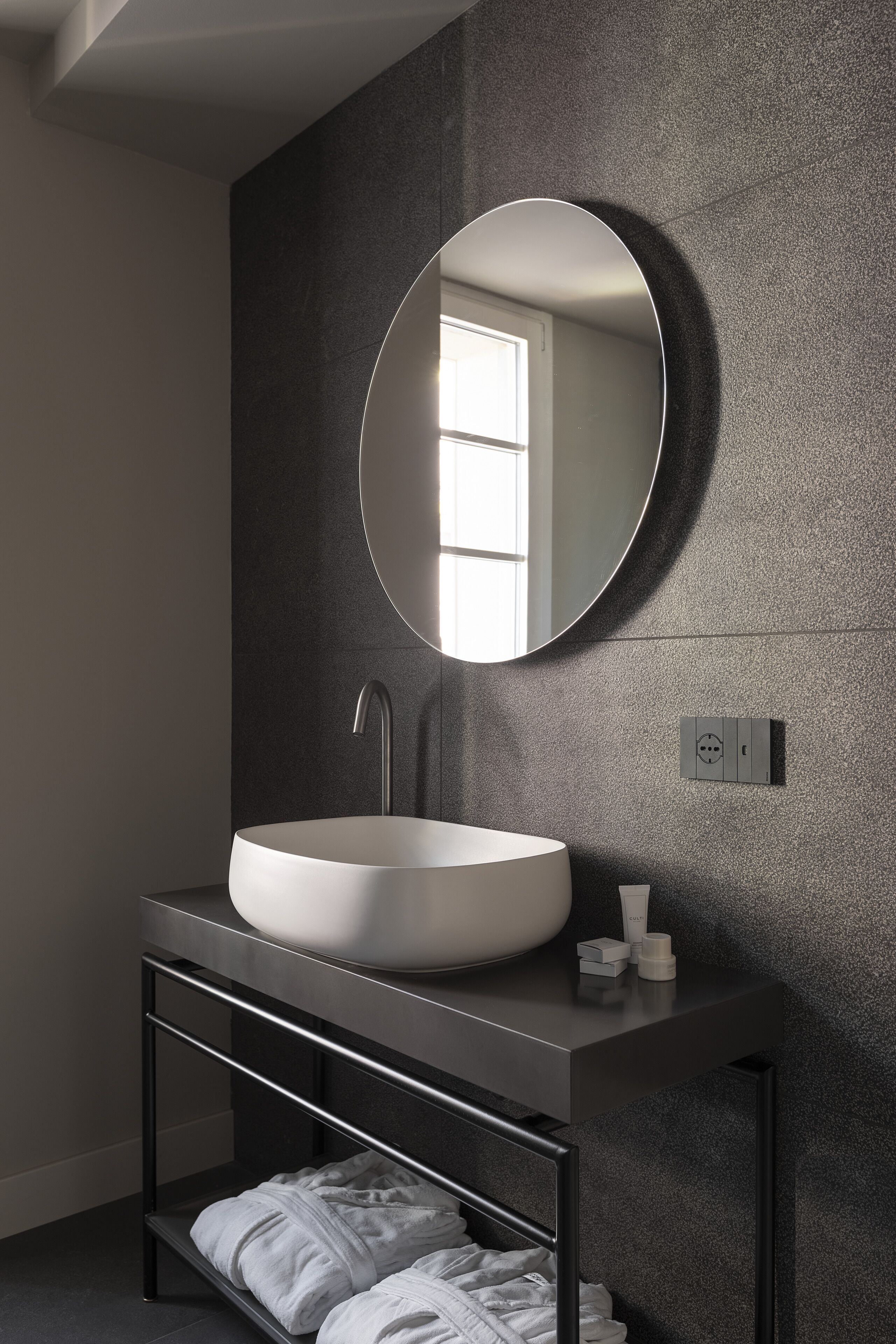 A modern bathroom with a round mirror, a white sink, and a shelf with towels.