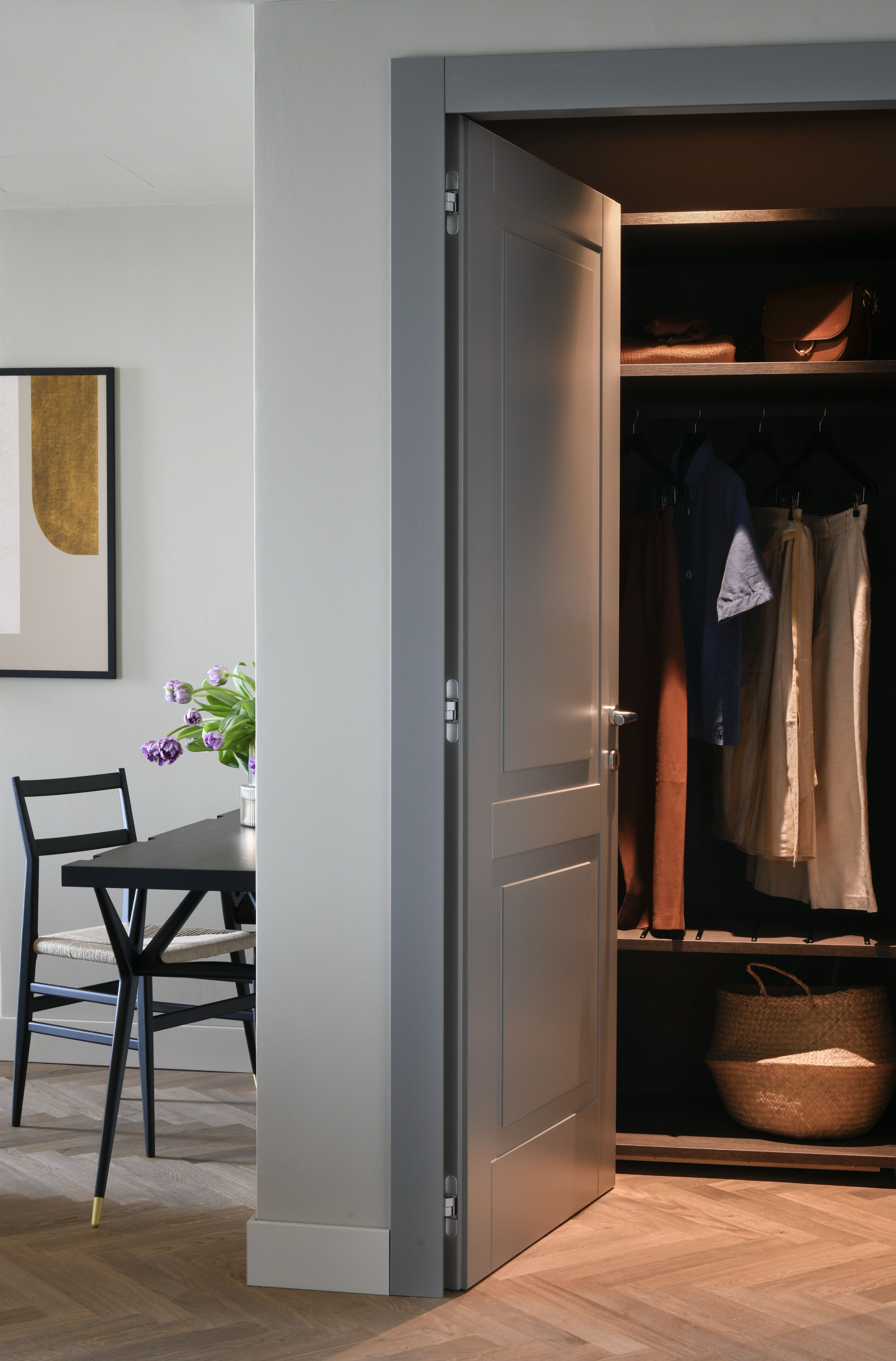 Quarto moderno com piso de madeira, mesa de jantar com cadeiras, um vaso com flores e um guarda-roupa com roupas penduradas e uma cesta.