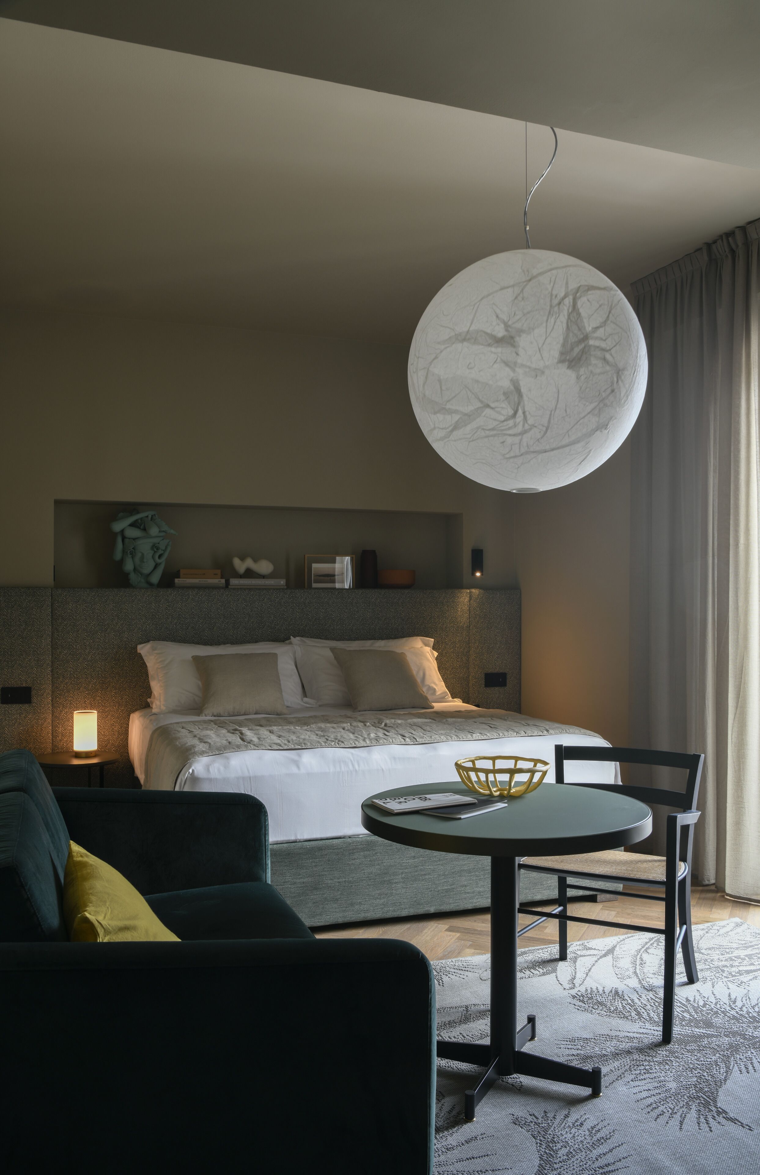 A modern hotel room with a bed, bedside table, lamp, round table, and chairs.