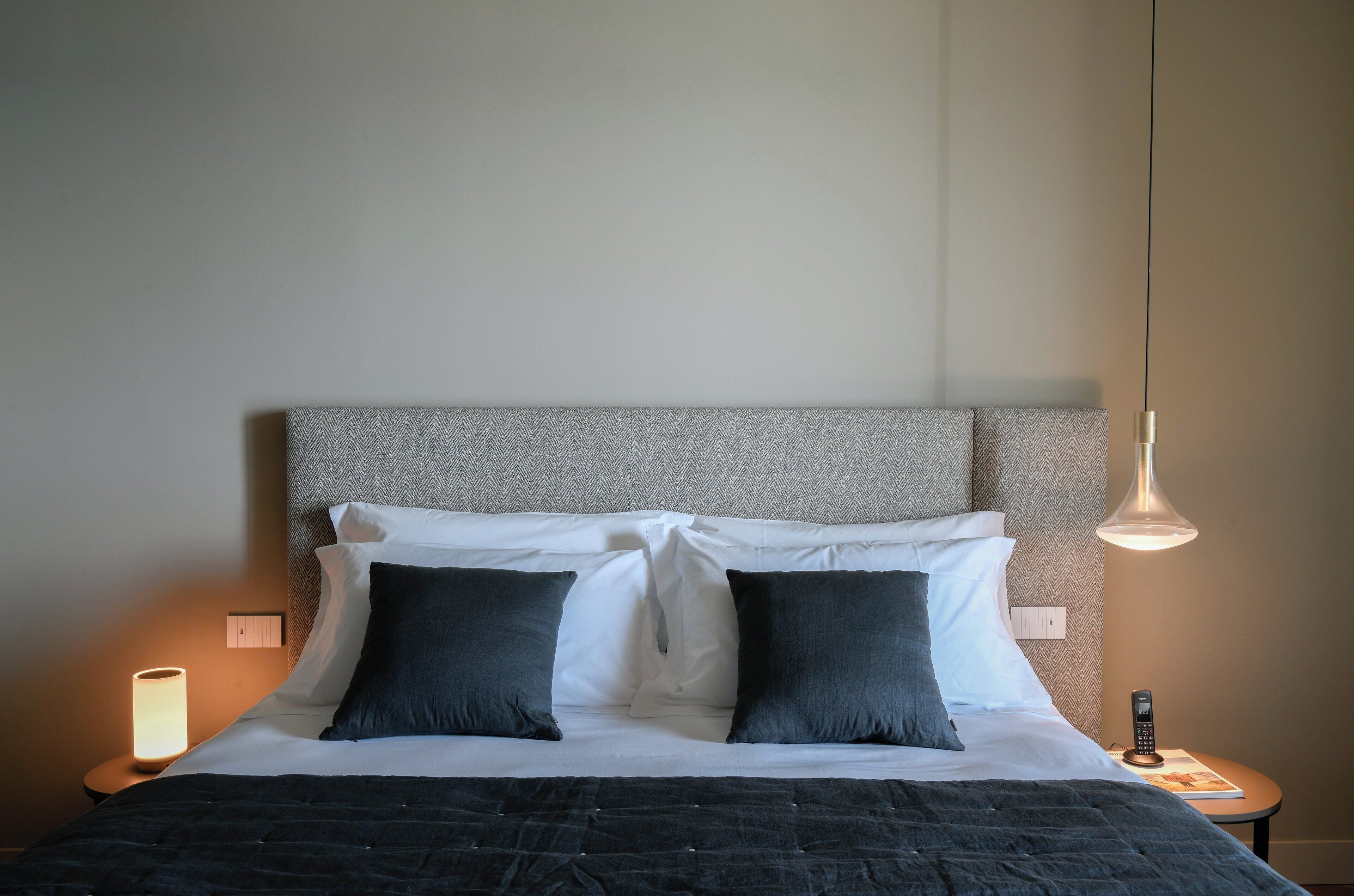 A neatly made bed with a grey headboard, two black pillows, and a white comforter. There is a bedside lamp on the left and a small table with a book and a remote control on the right.
