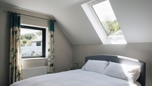 Een slaapkamer met dakraam, een bed met witte lakens, een nachtkastje met een lamp en gordijnen met een bladmotief.