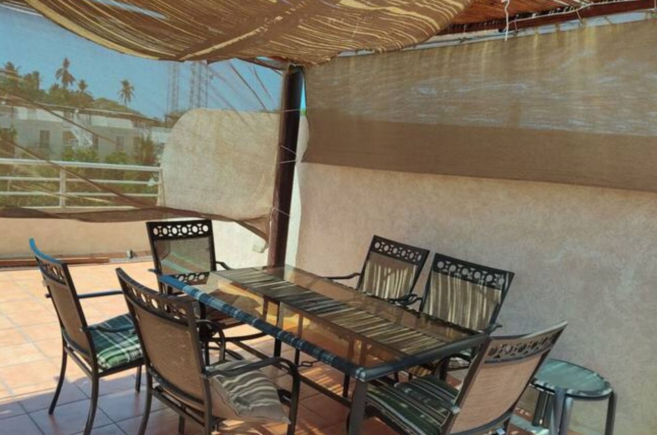 Un comedor con una mesa de vidrio y seis sillas, un amplio paraguas tejido y vistas a un edificio y palmeras.