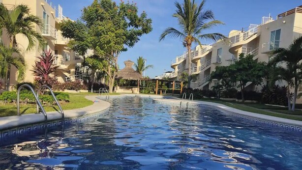 A swimming pool with a clear blue water surface, surrounded by palm trees and a building with balconies.