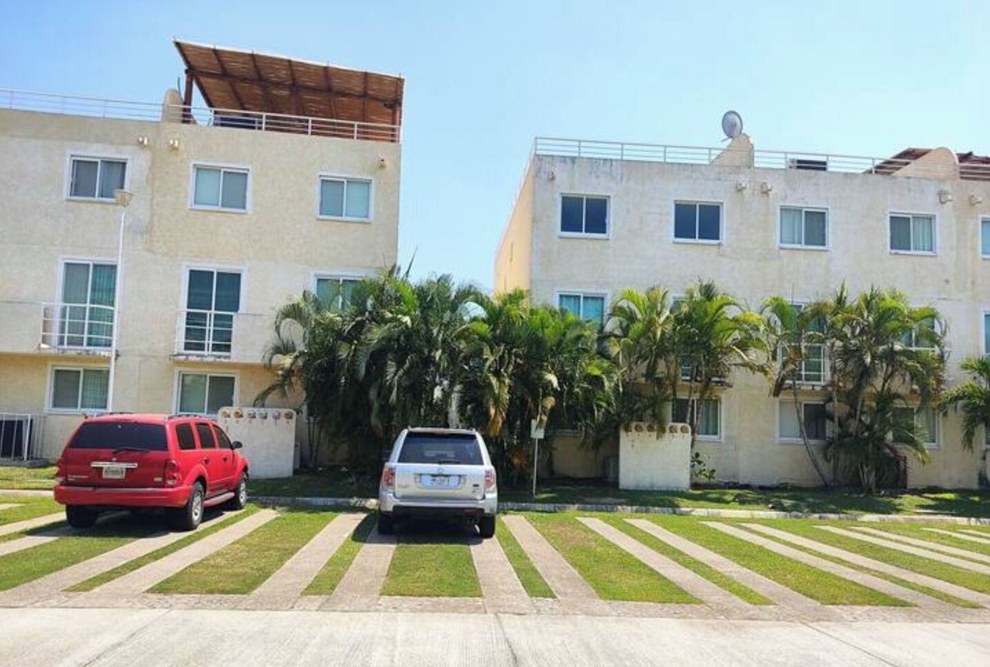 A residential building with multiple floors, a flat roof, and a satellite dish. There are two cars parked in the driveway, and the area is surrounded by palm trees.