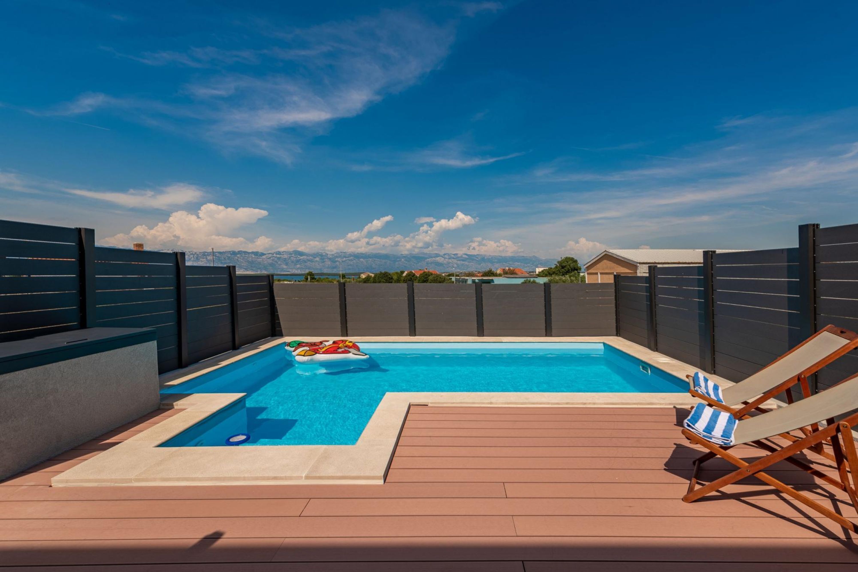 A swimming pool with a view of mountains, surrounded by a wooden deck and a fence.