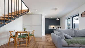 A modern living room with a grey sofa, wooden dining table, and chairs. A staircase with wooden steps and black railings leads to a kitchen area with dark cabinets and a white countertop. - Villa NiA Zadarvillas (Nin)
