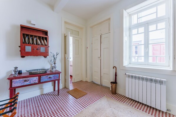 Una habitación con piso a cuadros, un radiador, una ventana con vista, una puerta y un armario con vajilla.