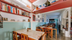 A dining area with wooden tables and chairs, a bookshelf with books, and a kitchen area in the background.