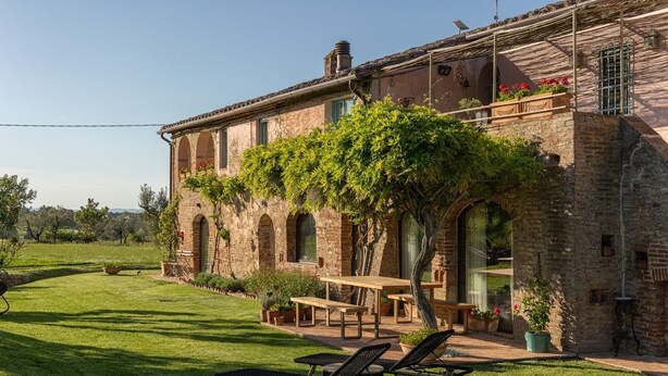 Un edificio rústico de piedra con un patio jardín, mobiliario exterior y una pérgola cubierta de plantas trepadoras.
