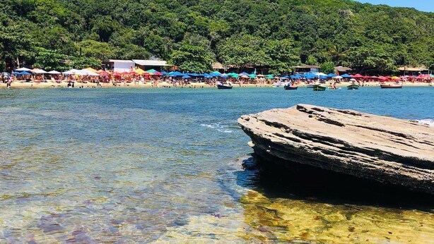 A clear beach with a rocky outcrop in the water, a forested hill in the background, and colorful umbrellas on the beach. - Pousada Porto Búzios (Búzios)