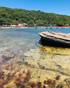 A clear beach with a rocky outcrop in the water, a forested hill in the background, and colorful umbrellas on the beach. - Pousada Porto Búzios (Búzios)