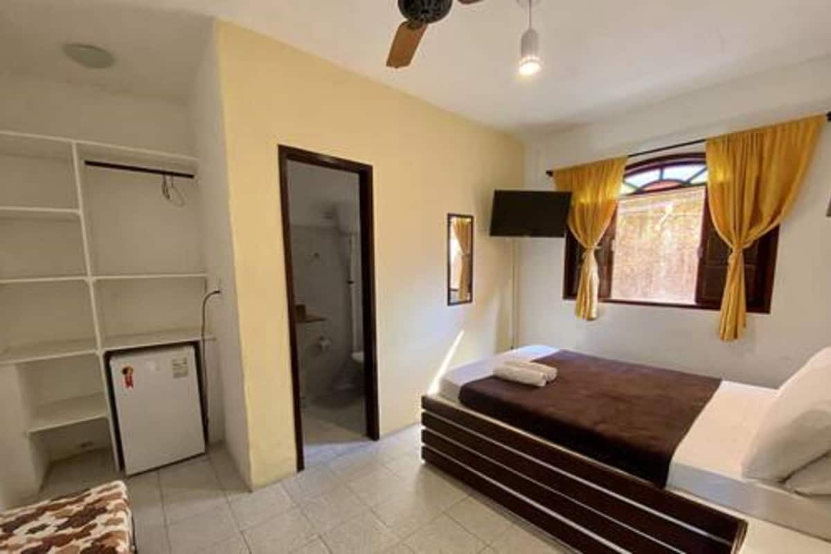 A bedroom with a bed, a wardrobe, a small fridge, a TV, and a window with curtains.