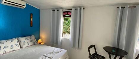 A bedroom with a bed, a desk, and chairs. There is a window with curtains, an air conditioning unit, and a lamp on the bedside table.
