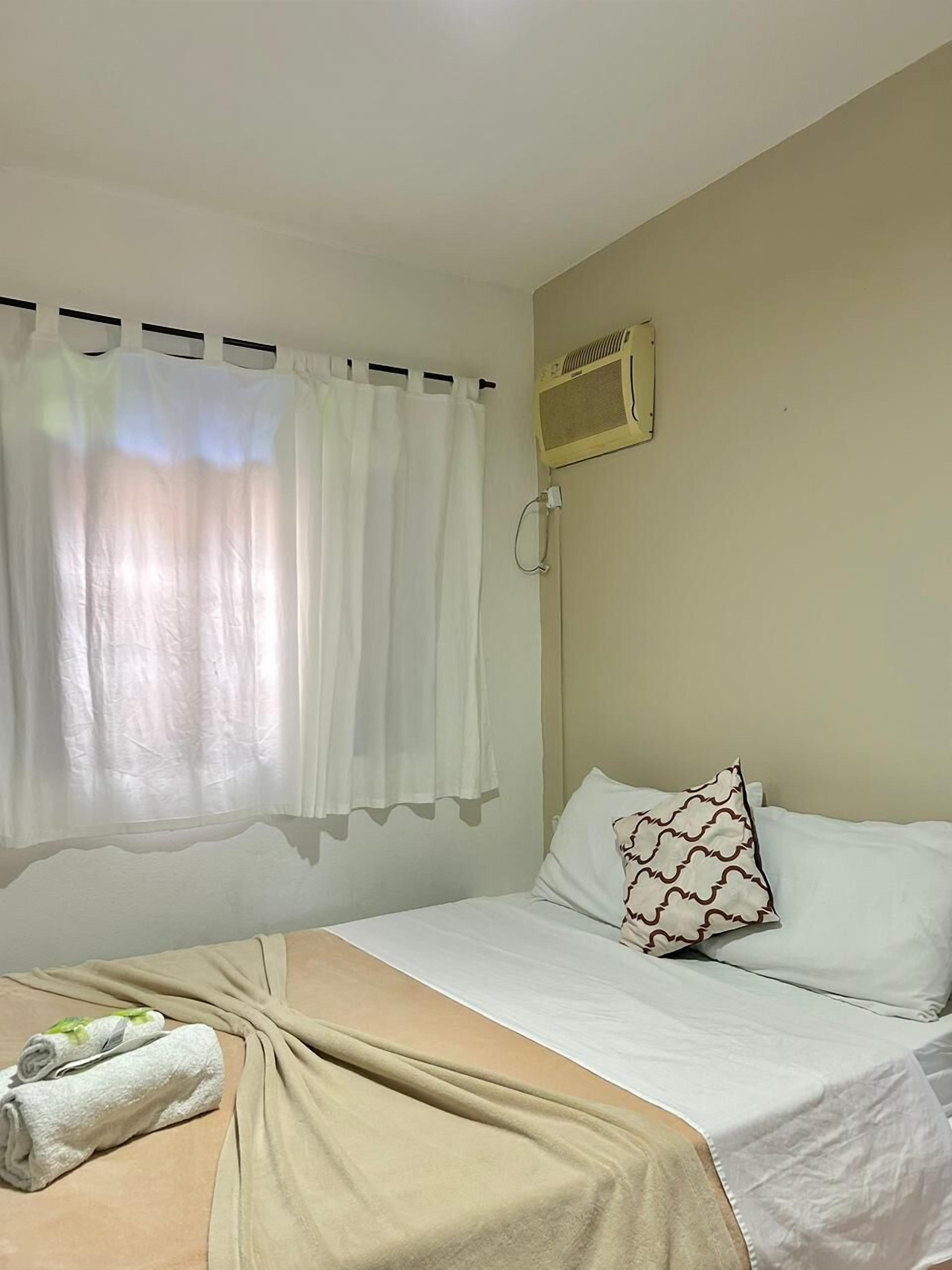 A single bed with a beige bedspread, white pillows, and a decorative pillow with a cloud pattern. A white towel is neatly folded on the bed.