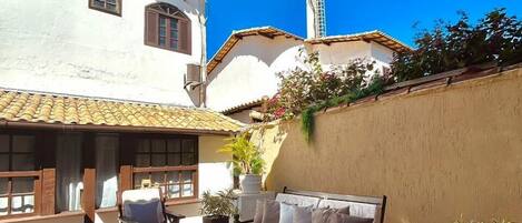 A courtyard with a white building, a tall tower, and outdoor seating with cushions and a coffee table.
