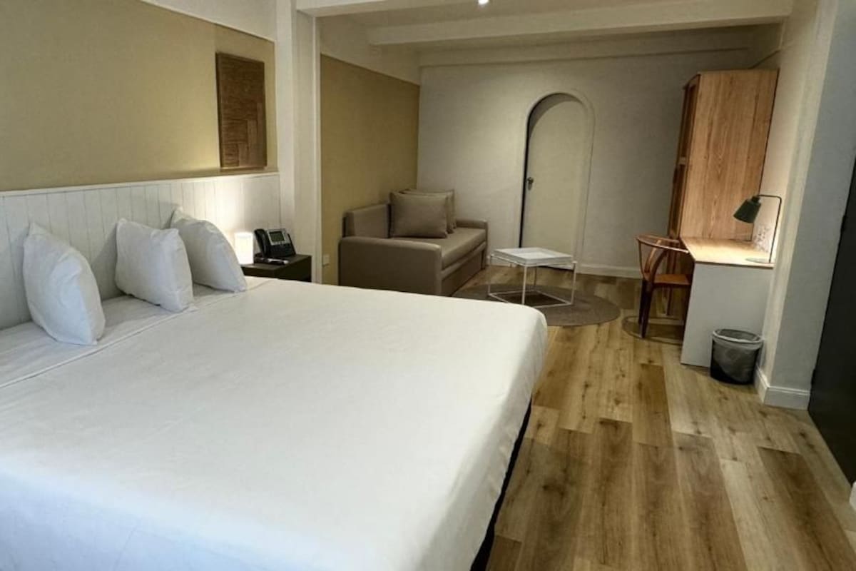 A hotel room with a large bed, a desk with a lamp, a chair, and a small table.
