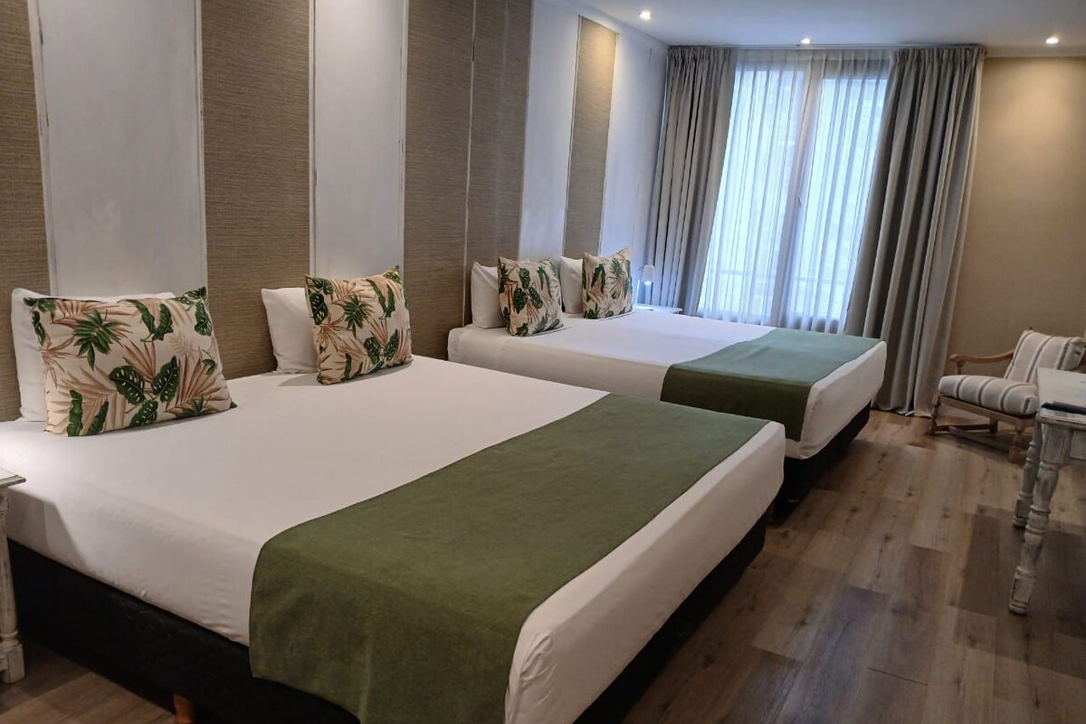 A hotel room with two beds, each with a green bedspread and tropical-themed pillows.