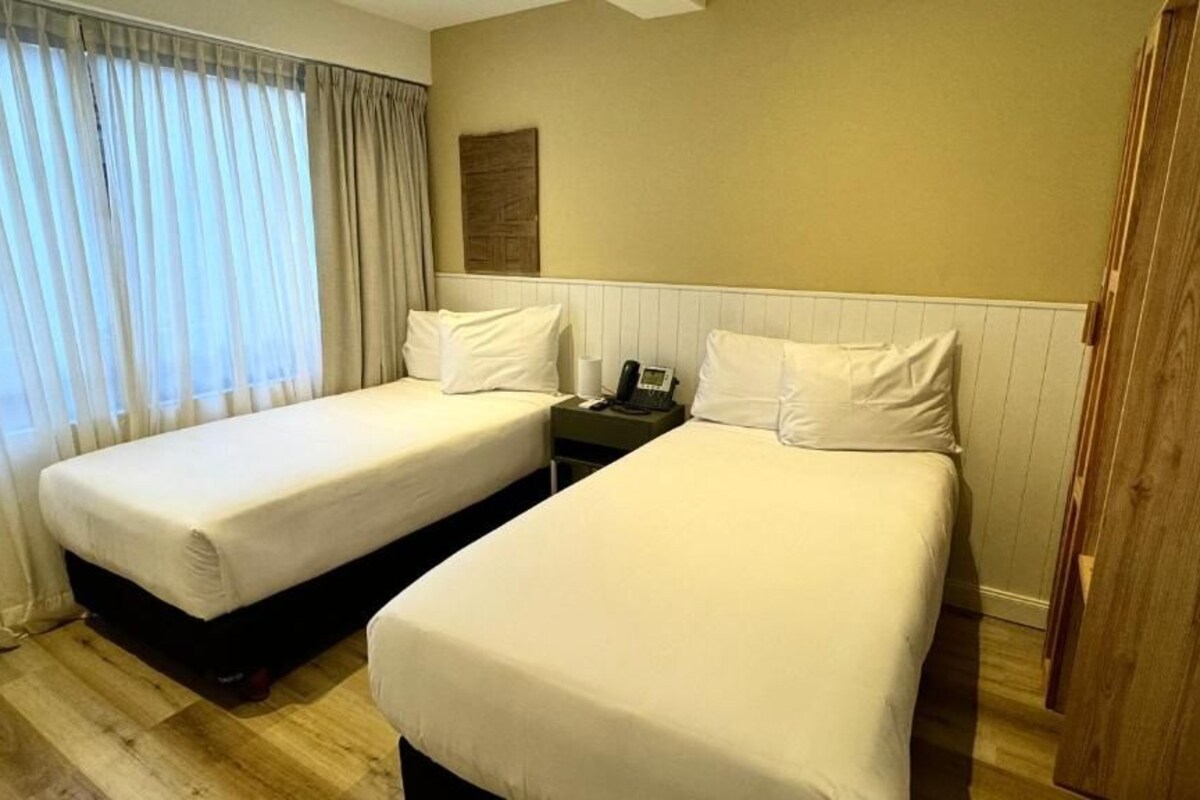 A hotel room with two single beds, a wooden floor, a bedside table with a phone, and a window with curtains.