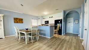 Una cocina moderna con armarios blancos, un refrigerador negro y una isla azul. Hay una zona de comedor con una mesa de madera y sillas blancas.