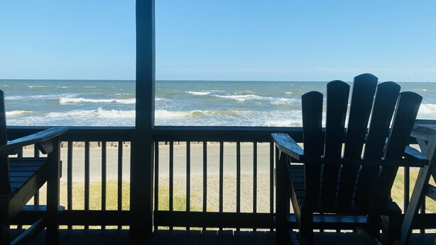 Una terraza de madera con dos sillas Adirondack mirando una playa con olas.