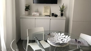 Moderni ruokailutila, jossa on lasipöytä, valkoiset tuolit, seinälle asennettu litteänäyttöinen televisio ja valkoinen kaappi sen alla.