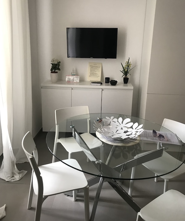 A modern dining area with a glass table, white chairs, a flat-screen TV mounted on the wall, and a white cabinet below.