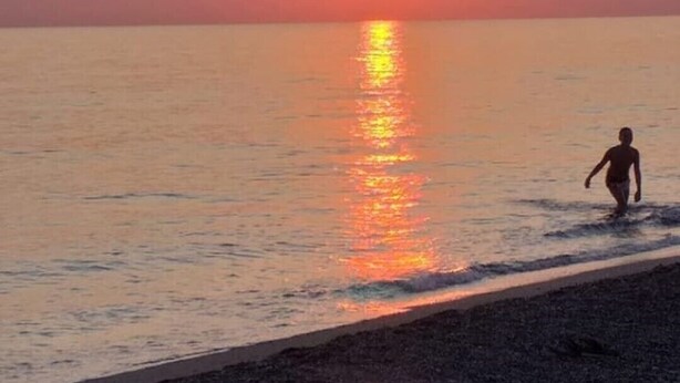 Rantamaisema auringonlaskun aikaan, tyyni vesi ja henkilö kävelemässä rantaa pitkin.