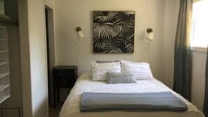 A neatly made bed with white bedding, a gray throw, and a pillow with the text "live in the moment." There is a wall-mounted painting of palm leaves, two bedside lamps, and a window with curtains. - Enjoy the Summertime Beaches, Concerts and Events in Prince Edward County! (Prince Edward)
