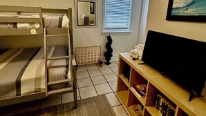 Una habitación con literas, un televisor, una estantería, una ventana con persianas y una pequeña planta decorativa.