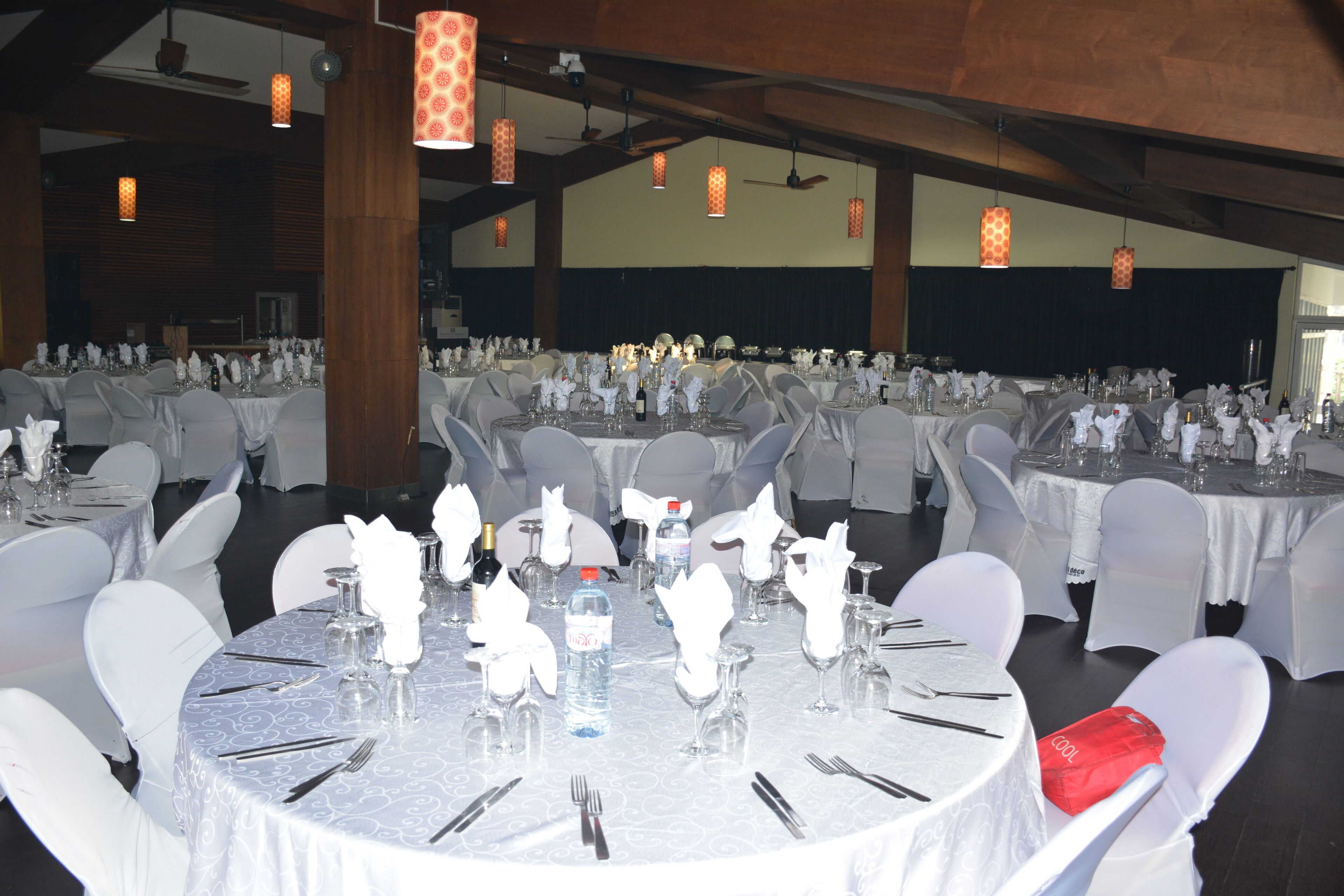 Un salón de banquetes con mesas redondas dispuestas para un evento formal, con manteles blancos, cristalería y sillas cubiertas de blanco.