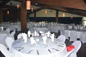 Un salón de banquetes con mesas redondas dispuestas para un evento formal, con manteles blancos, cristalería y sillas cubiertas de blanco.