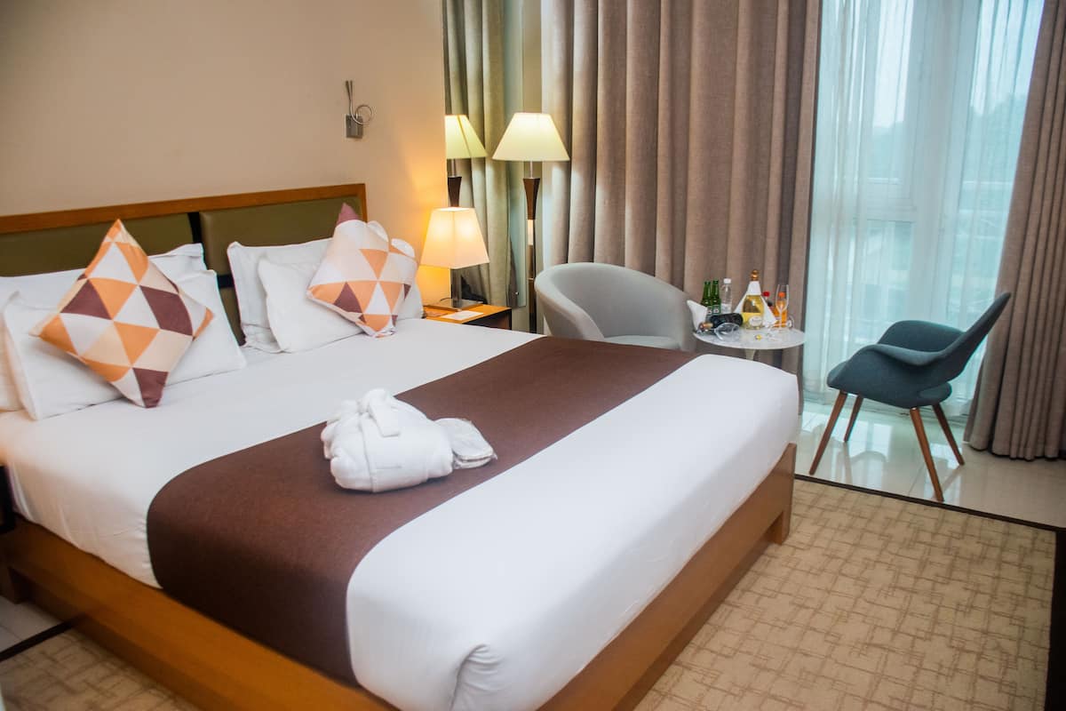 A hotel room with a large bed, a desk with a chair, a small table with drinks, and a view of the city through the window.