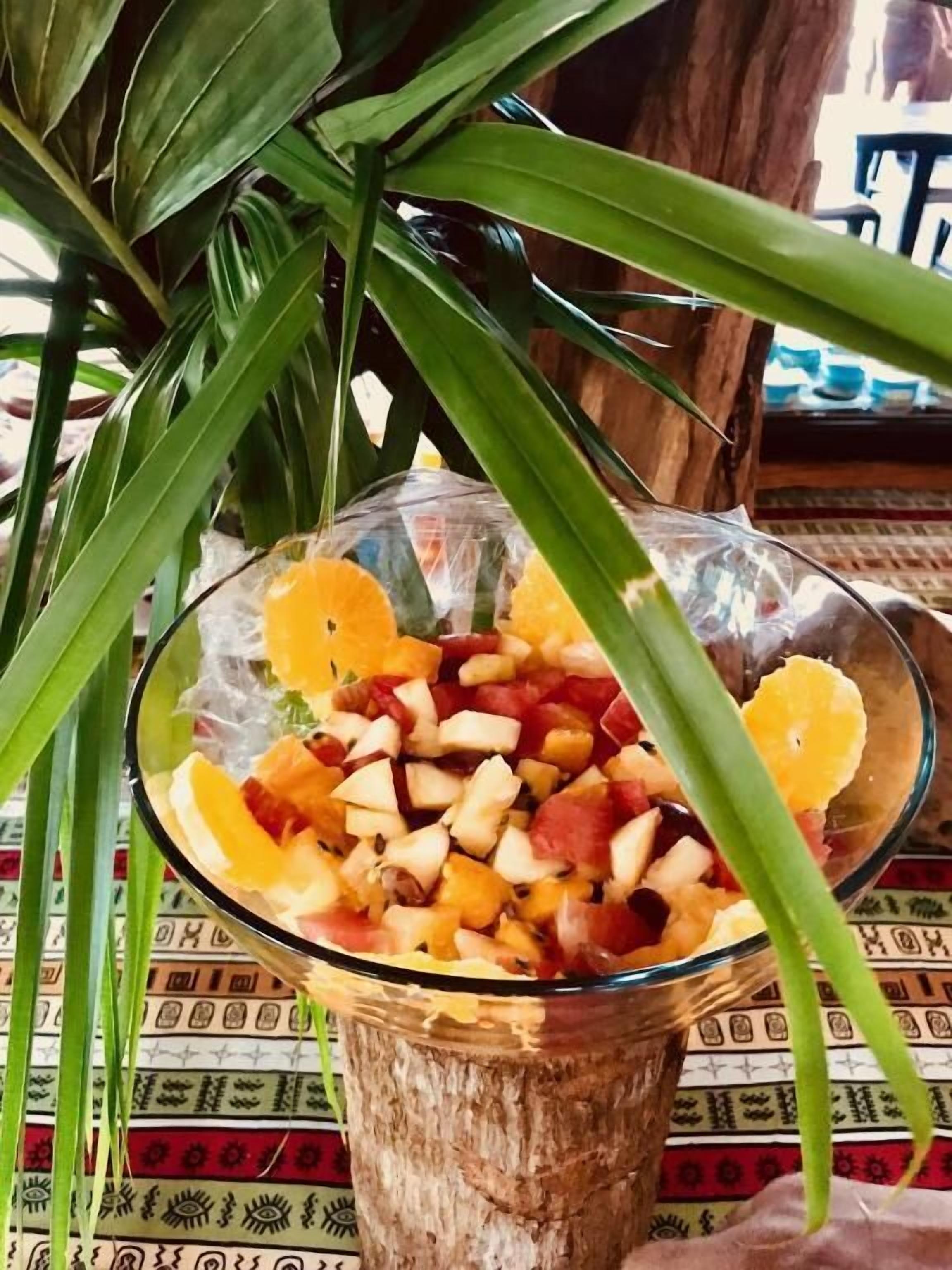 A glass bowl filled with assorted fruits on a wooden table with a patterned tablecloth.