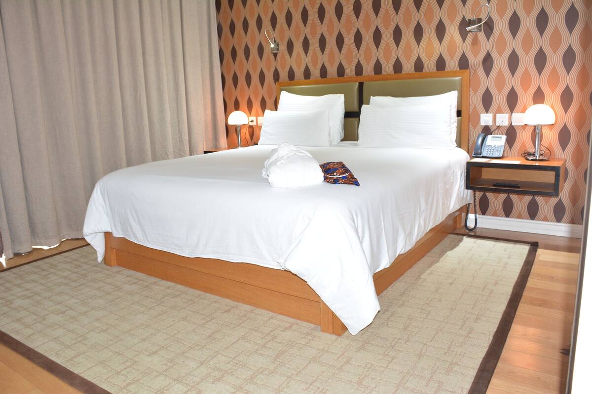 A hotel room with a large bed, two bedside lamps, a nightstand with a phone, and a patterned wall.
