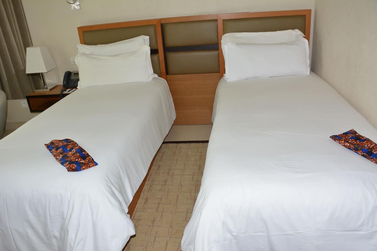 A hotel room with two single beds, a nightstand with a lamp and phone, and a carpeted floor.