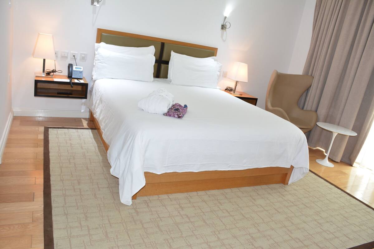 A hotel room with a large bed, a wooden headboard, a nightstand with a lamp and phone, a chair, and a small table.