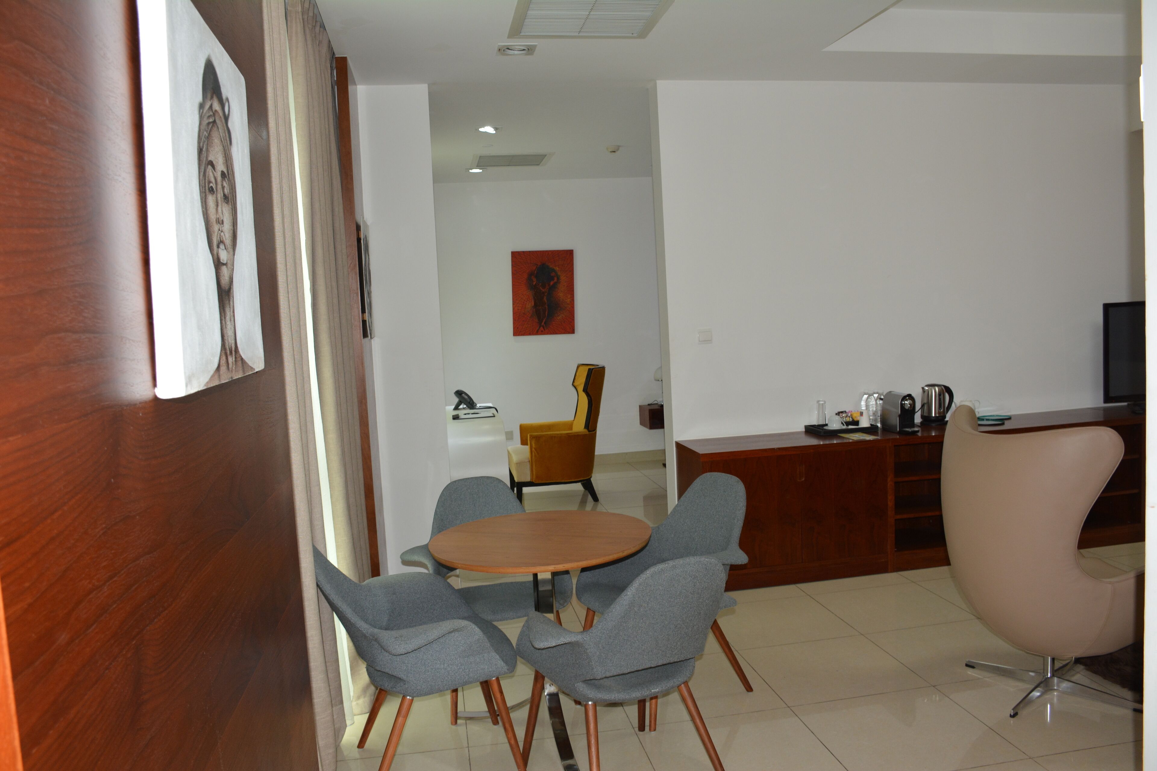 A modern dining area with a wooden table and grey chairs, a wooden cabinet with kitchen appliances, and a yellow chair in the background.