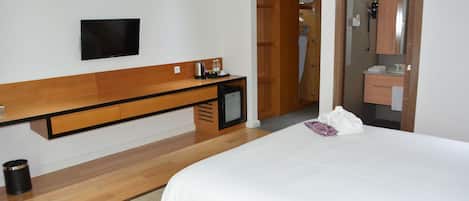 A hotel room with a bed, a TV mounted on the wall, a wooden desk, a small refrigerator, and a bathroom with a mirror and sink.