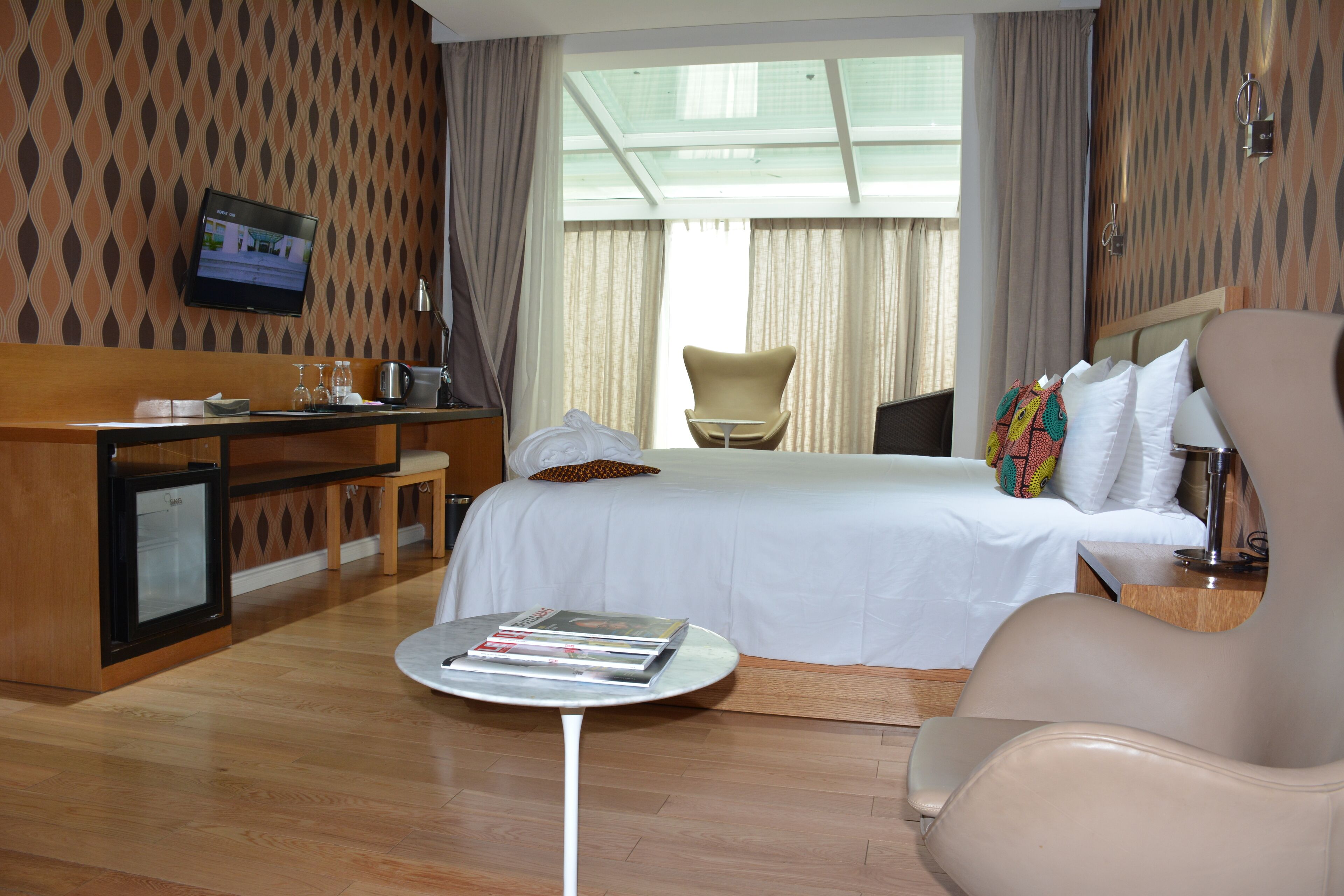 A hotel room with a bed, a desk with a computer, a chair, a small table with magazines, and a TV on the wall.